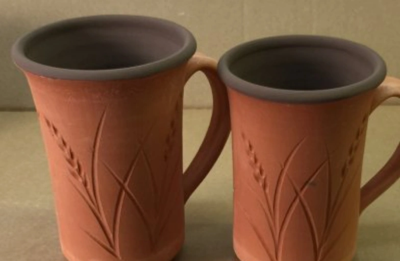 Two terracotta mugs with intricate leaf patterns and smooth gray rims placed side by side on a wooden surface.