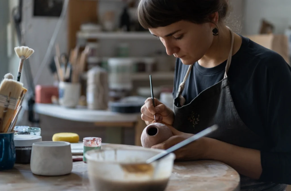 Woman concentrating while hand-painting a small ceramic vase in a pottery studio. Creative workspace with brushes and pottery tools in the background.