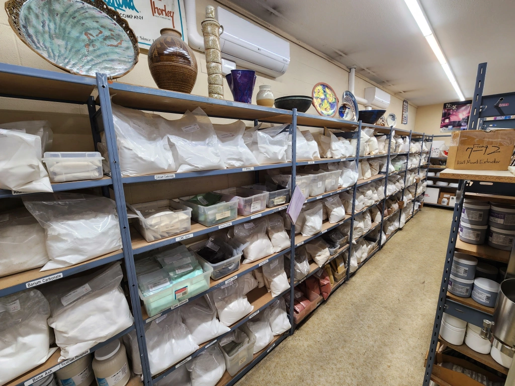 Shelves stocked with numerous white bags of pottery clay and various ceramic pieces in a pottery studio or supply room.