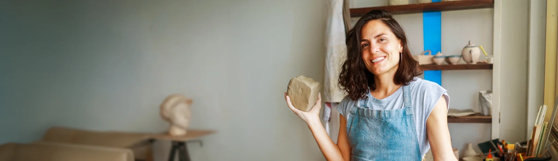 Woman in a blue apron holding a ceramic piece in a pottery studio, surrounded by shelves with various clay creations.