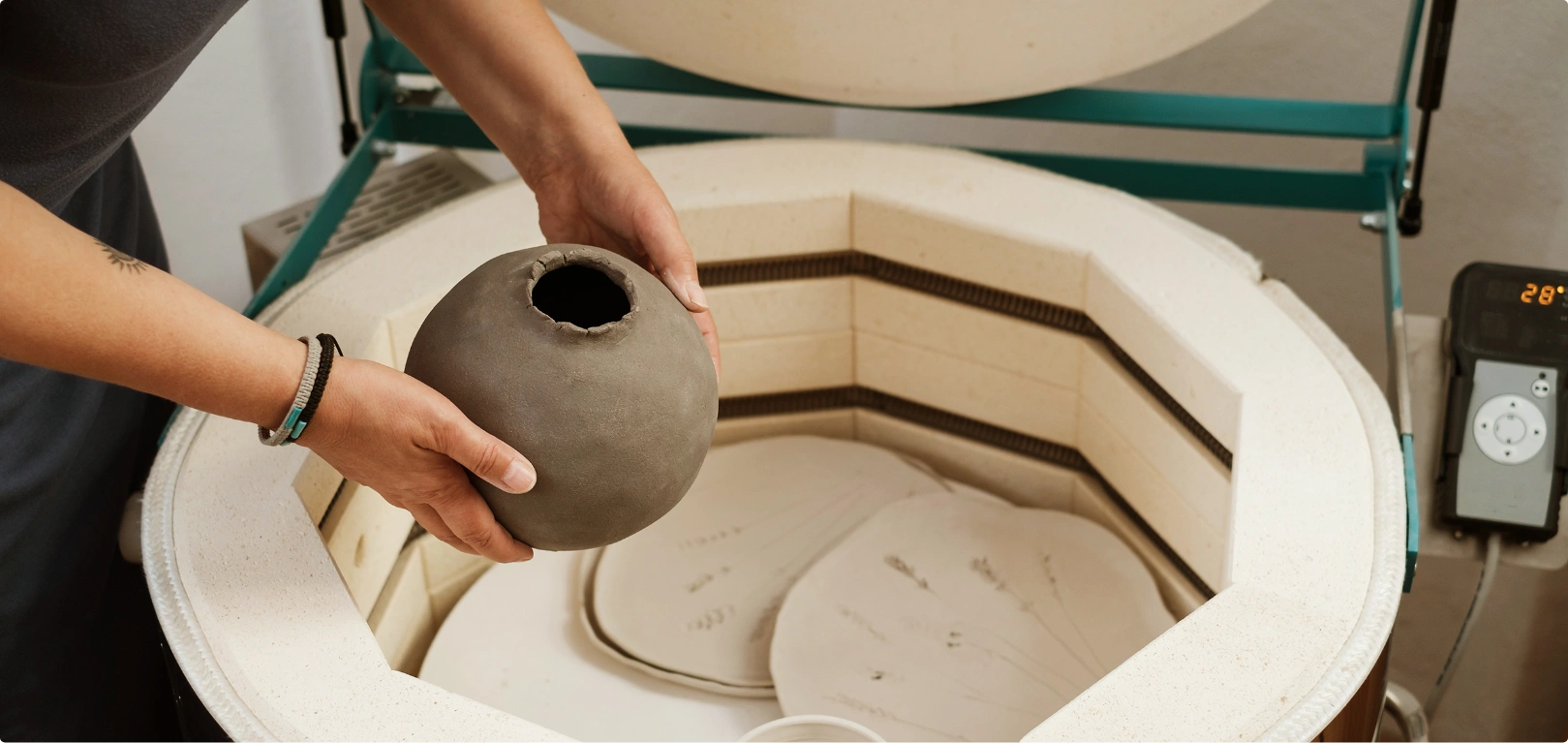 Hands placing a round clay pot inside a pottery kiln ready for firing.