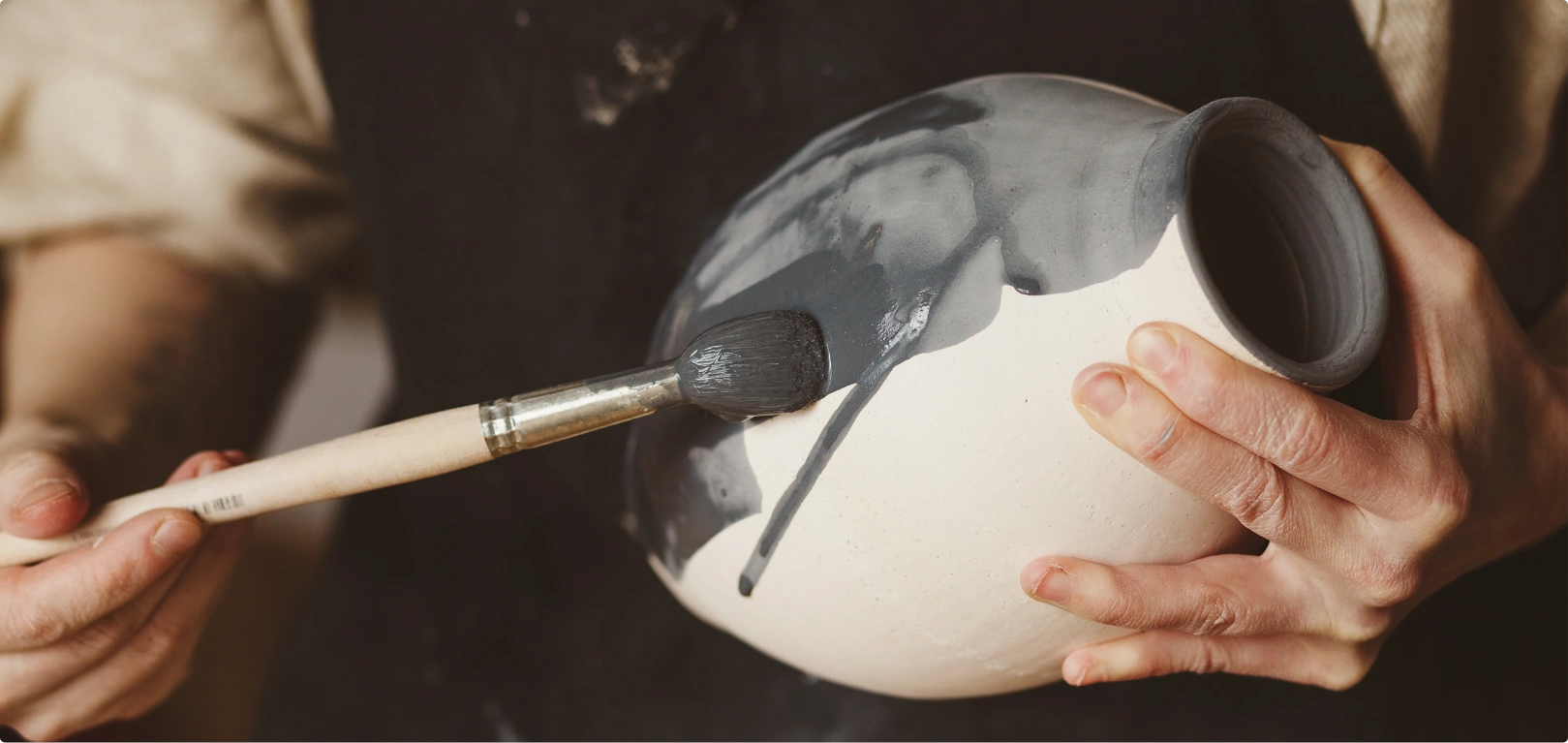 Close-up of hands painting detailed designs on a ceramic pot with a fine brush.
