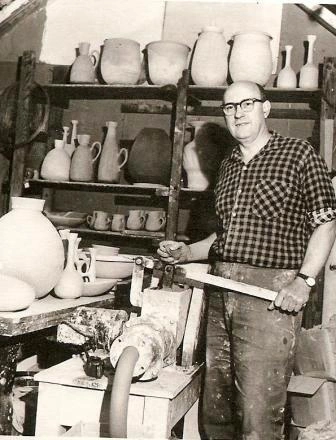 Black and white photo of a man shaping pottery on a wheel in a workshop filled with various ceramic pots and jugs on shelves.